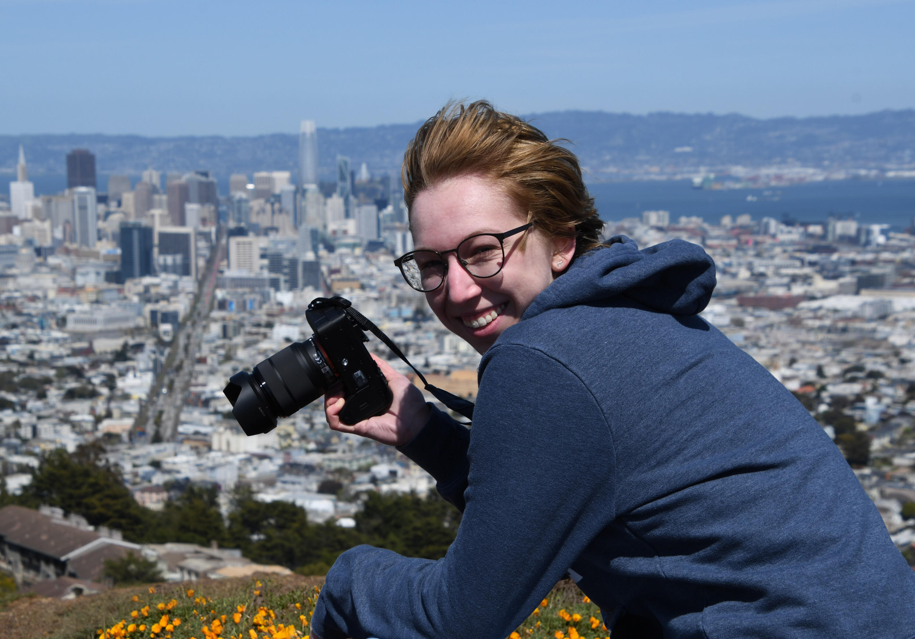 A photo shows a smiling Natalie Gospe as the hold a camera to take photos over the San Francisco skyline. There are California Poppies in bloom below them.